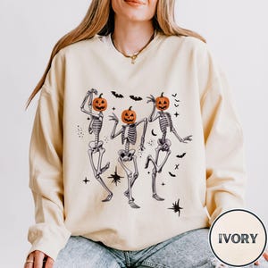 May include: A cream colored sweatshirt with a graphic of three skeletons wearing pumpkin heads. The skeletons are dancing and there are bats and stars around them. The text "IVORY" is in the bottom right corner of the image.