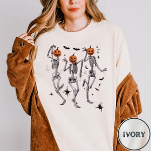 May include: A white long-sleeved shirt with a graphic of three skeletons dressed as pumpkins dancing. The shirt is labeled "IVORY" in the bottom right corner.