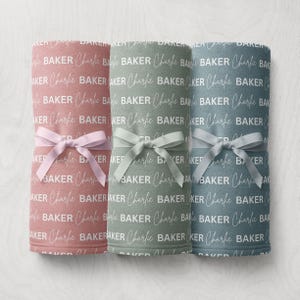 May include: Three rolled baby blankets in pink, green, and blue, each tied with a matching ribbon. The blankets are printed with the name "Charlie Baker" in a repeating pattern. A soft and cozy baby item.