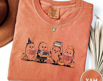 May include: A burnt orange t-shirt with a graphic of four cartoon yams dressed as witches and holding books. The text "COMFORT COLORS" is on the tag. The shirt says "YAM" in the bottom right corner.