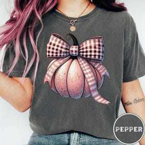 Puede incluir: Una camiseta gris con una calabaza rosa con un lazo de cuadros rosa y blanco. La calabaza está centrada en la camiseta.