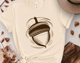 Puede incluir: Una camiseta beige con un gráfico de bellota marrón. La bellota está estilizada con un diseño de remolino a su alrededor.