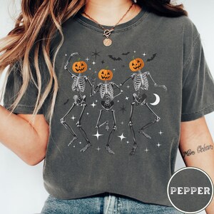 May include: A dark gray t-shirt with a graphic of three skeletons dancing. Each skeleton has a pumpkin head with a jack-o-lantern face. The graphic is surrounded by stars and bats.