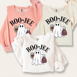 May include: Three white sweatshirts with a ghost holding a coffee cup and a purse. The text "BOO-JEE" is printed on each sweatshirt in black with a distressed look. The sweatshirts are in different colours: pink, beige, and white.