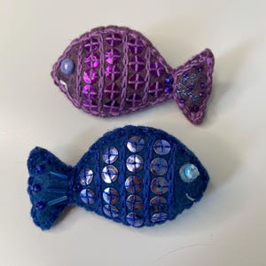 Peut inclure: Deux ornements de poisson en feutre faits à la main. L'un est violet avec des paillettes roses et un œil bouton. L'autre est bleu avec des paillettes argentées et un œil en perle bleue. Les deux ont des détails brodés et un design fantaisiste.