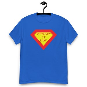 Könnte beinhalten: Königsblaues T-Shirt mit einem rot-gelben rautenförmigen Aufdruck. Der Aufdruck zeigt die Worte "ATTORNEY AT LAW" in einer fetten, serifenlosen Schrift. Das T-Shirt hängt an einem schwarzen Kleiderbügel.