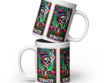 May include: Two white ceramic mugs with a colorful tarot card design featuring a skeleton smoking a cigarette. The design includes the text "THE STONER" and cannabis leaves. The mugs are stacked on top of each other.