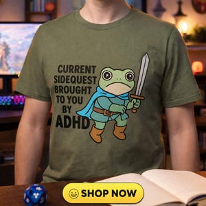 May include: Olive green t-shirt featuring a cartoon frog warrior holding a sword. The text on the shirt reads "CURRENT SIDEQUEST BROUGHT TO YOU BY ADHD". A "SHOP NOW" button is visible at the bottom.