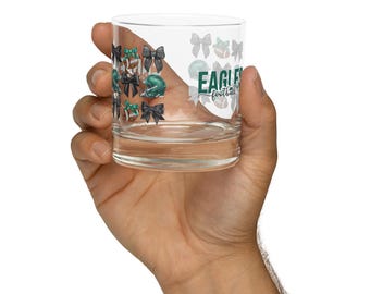May include: A clear glass tumbler with a design featuring footballs, helmets, and bows in green, black, and white. The words "Eagles football" are printed on the side of the glass.