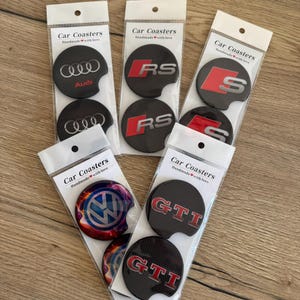 Peut inclure: Plusieurs dessous de verre pour voiture dans un emballage plastique transparent. Ils présentent des logos de marques automobiles comme Audi, RS, S, GTI et Volkswagen. Les dessous de verre sont noirs avec des logos argentés, rouges et blancs. L'emballage indique "Car Coasters Handmade with love."