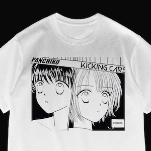 Może przedstawiać: Biała koszulka z czarno-białą grafiką w stylu mangi. Grafika zawiera dwie postacie w stylu anime i tekst "PANCHIKO KICKING CARS" nad nimi, ze słowem "SHOCKED" poniżej.