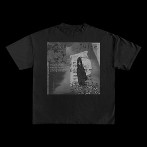 Op de afbeelding: Zwart t-shirt met een grijswaarden grafisch ontwerp. Het ontwerp bevat een illustratie van een persoon, het woord "julie" en de herhaalde zin "pushing daisies". Extra tekst luidt "CONFIRMATION OF ENTRY DISPATCH".