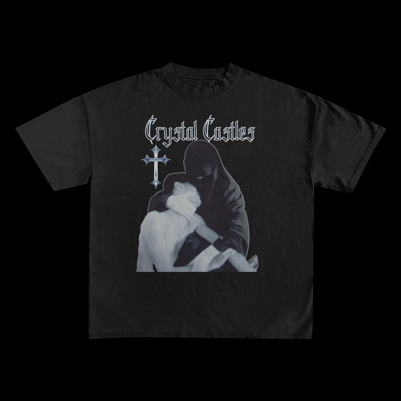 Puede incluir: Camiseta negra con el nombre de la banda "Crystal Castles" en una fuente estilizada. El dise&ntilde;o muestra dos figuras, una con una capucha negra, abrazando a otra. Tambi&eacute;n hay una cruz.