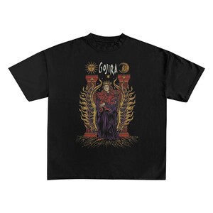 May include: Black t-shirt featuring a graphic of a robed figure with a crown, holding a snake, and surrounded by flames. The word "GOJIRA" is above the figure, with sun and moon symbols on either side. The design is in shades of red, purple, and gold.