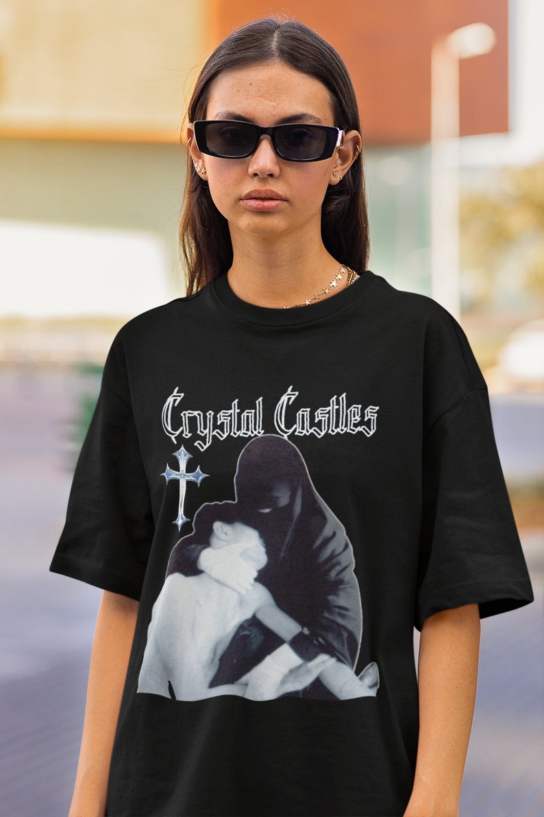 Puede incluir: Camiseta negra con el nombre de la banda "Crystal Castles" en fuente g&oacute;tica blanca sobre un gr&aacute;fico en blanco y negro. El gr&aacute;fico representa a una persona con capucha negra abrazando a otra persona. La cruz es de color azul claro.