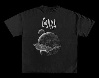 May include: Black t-shirt featuring a white graphic of a whale swimming around a planet with the word "GOJIRA" above. The design is a detailed illustration with a celestial theme.