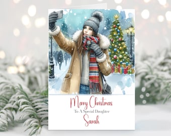 Puede incluir: Tarjeta de Navidad con una ilustración en acuarela de una persona haciéndose un selfie en una escena invernal nevada. La persona lleva abrigo, gorro y bufanda. La tarjeta dice "Feliz Navidad A Una Hija Especial Sarah."