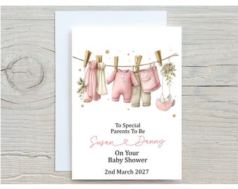 Puede incluir: Tarjeta de baby shower con una ilustración en acuarela de ropa de bebé colgada en un tendedero. La tarjeta dice "To Special Parents To Be Susan & Danny On Your Baby Shower 2nd March 2027". La tarjeta es blanca con un sobre blanco.