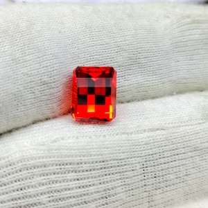 Könnte beinhalten: Ein facettierter, rechteckig geschliffener, roter Edelstein. Der Stein hat ein Schachbrettmuster im Inneren, mit roten und schwarzen Quadraten. Der Stein liegt auf einem weißen, strukturierten Hintergrund.