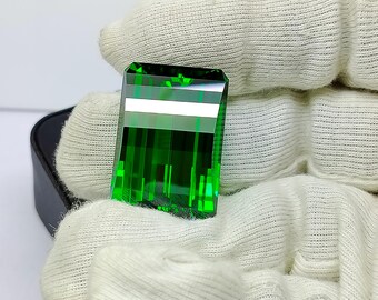 May include: A rectangular, emerald-green gemstone with a faceted cut. The gem is held in a white glove, showcasing its clarity and vibrant color. The gem's facets reflect light, creating a sparkling effect.