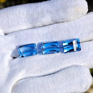 May include: Three rectangular, light blue gemstones are displayed. The gems have a vibrant, transparent appearance, with visible internal structures. The gems are arranged horizontally, showcasing their polished surfaces and colour. The gemstones are displayed on a white surface.