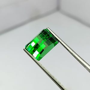 May include: A vibrant, emerald-cut green gemstone is held by silver tweezers. The gem's facets reflect light, showcasing its clarity and brilliance. The rectangular shape and intense color suggest a precious stone, possibly for jewelry.
