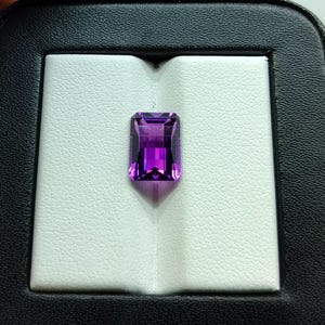 Puede incluir: Una gema de amatista rectangular y facetada con un tono púrpura intenso. La gema se presenta sobre un fondo blanco texturizado dentro de un estuche negro, mostrando su claridad y color.