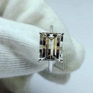 May include: A silver ring featuring a large, clear, rectangular emerald-cut gemstone. The gemstone has a subtle brown hue.