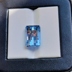 May include: A rectangular, light blue gemstone with a faceted surface, presented in a black jewellery box with a white interior. The gem's colour is a vibrant, clear blue, reflecting light. The box has a dark blue, textured exterior.