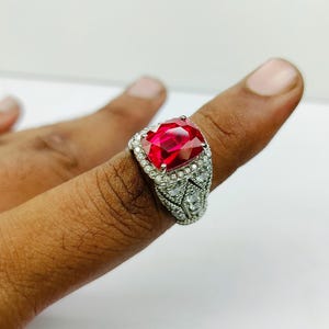 May include: A silver-toned ring featuring a large, faceted, red gemstone. The ring is adorned with smaller, clear gemstones around the main stone and along the band, creating a sparkling effect. The ring is worn on a finger.