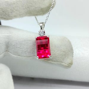 Puede incluir: Un collar con colgante de plata con una gema rectangular de color rojo vibrante. Una pequeña piedra redonda y transparente se encuentra encima de la piedra roja más grande. El collar se muestra sobre un fondo blanco.