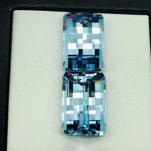 May include: Two light blue aquamarine gemstones in a square emerald cut, displayed in a white box with a black border.