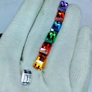 May include: A selection of vibrant, rectangular gemstones in a vertical arrangement, displaying a rainbow of colours from purple to yellow. A separate, clear gemstone is also visible. The gems are held against a white fabric background.