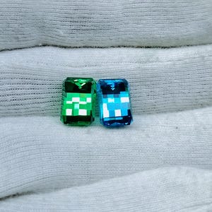 May include: Two rectangular gemstones, one green and one blue, with a chequered pattern. The gemstones are displayed on a white fabric surface.