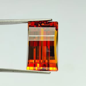 以下が含まれることがあります： 赤みがかったオレンジ色の長方形のファセットカットされた宝石。宝石はピンセットで保持されています。