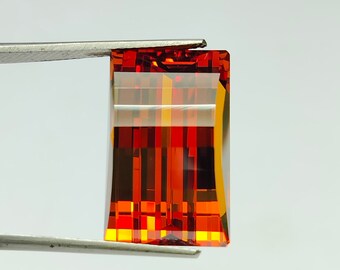 May include: A rectangular, faceted gemstone with a reddish-orange hue. The gemstone is held in place by a pair of tweezers.