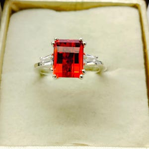 May include: A silver ring with a large, red, emerald-cut gemstone and two smaller, clear, baguette-cut gemstones on the sides.