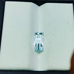 May include: A rectangular, aquamarine-colored gemstone with a step-cut design. The gem is set against a white, textured background within a black box, showcasing its clarity and brilliance. The gem's facets reflect light, creating a sparkling effect.
