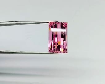 May include: A pink rectangular gemstone with a faceted cut, held by tweezers against a white background.