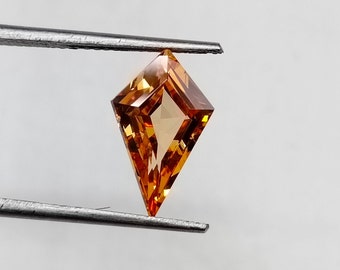 May include: A faceted, orange-brown gemstone with a kite shape, held by tweezers against a white background.