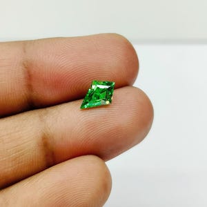 May include: A vibrant, emerald-green gemstone with a unique kite-like shape. The gem is held between fingers, showcasing its facets and brilliant color. The gemstone's cut allows for maximum light reflection, creating a sparkling effect. This gemstone is ideal for jewelry.