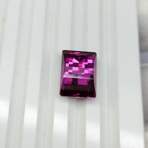 Può includere: Una gemma rettangolare sfaccettata di un vivido colore magenta-rosa. La pietra presenta una superficie chiara e lucida con bordi geometrici netti e un taglio a faccette multiple che crea un motivo a griglia di riflessi luminosi.