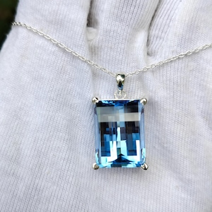 Puede incluir: Un collar de plata con un colgante rectangular de piedra preciosa azul claro. El colgante está engastado en un marco de plata y cuelga de una delicada cadena de plata. La piedra preciosa tiene un aspecto facetado.