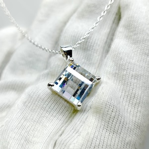 May include: A square-cut, clear crystal pendant necklace with a silver chain. The crystal is faceted, reflecting light. The pendant is set in a silver bezel with small silver accents at each corner. The necklace is displayed on a white surface.