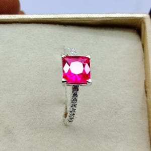 May include: A silver ring featuring a large, square-shaped, bright red gemstone surrounded by smaller, clear gemstones.