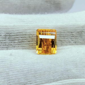 May include: A rectangular, faceted, golden-yellow gemstone. The gem has a bright, reflective surface with internal lines and a white band across the top. The stone is set against a white, textured background.