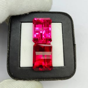 May include: Two rectangular, faceted, hot pink gemstones are displayed in a black tray with a white interior. The gems are stacked vertically, showcasing their vibrant colour and intricate cuts. The gems are held by a gloved hand.