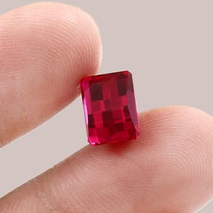 May include: A rectangular-cut ruby gemstone, displaying a rich, deep red hue. The gem is held between fingers, revealing its facets and polished surface. The background is a neutral tone, highlighting the ruby's colour and clarity.