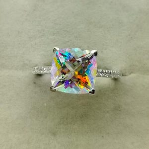 May include: A silver ring with a large, square-cut, iridescent gemstone. The gemstone displays a spectrum of colors, including orange, yellow, green, and blue. The band is adorned with small, clear stones. The ring is set against a neutral background.