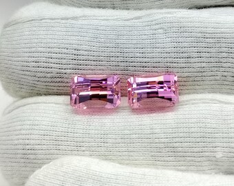May include: Two pink rectangular gemstones with a faceted cut. The gemstones are displayed on a white fabric background.